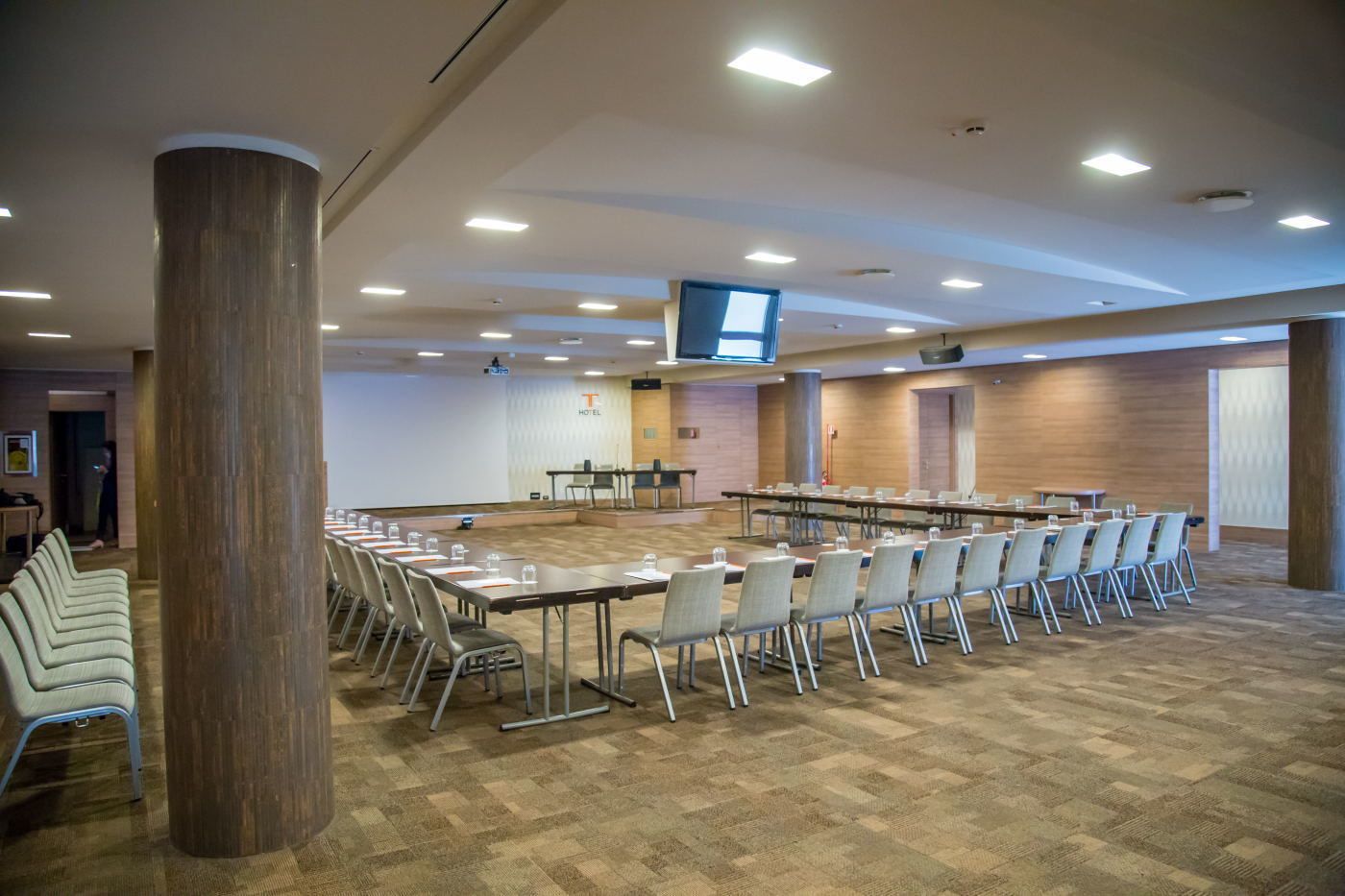 Una sala conferenze con diverse tavole e sedie, un ampio pilastro e uno schermo televisivo a soffitto.