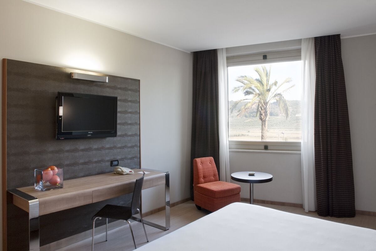 Habitación de hotel con cama, escritorio con silla, televisión, mesita y ventana con cortinas.