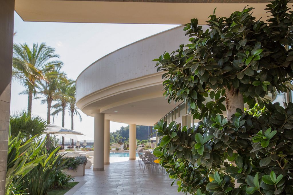 A modern hotel with a curved roof structure, a pool area with lounge chairs, and lush greenery.