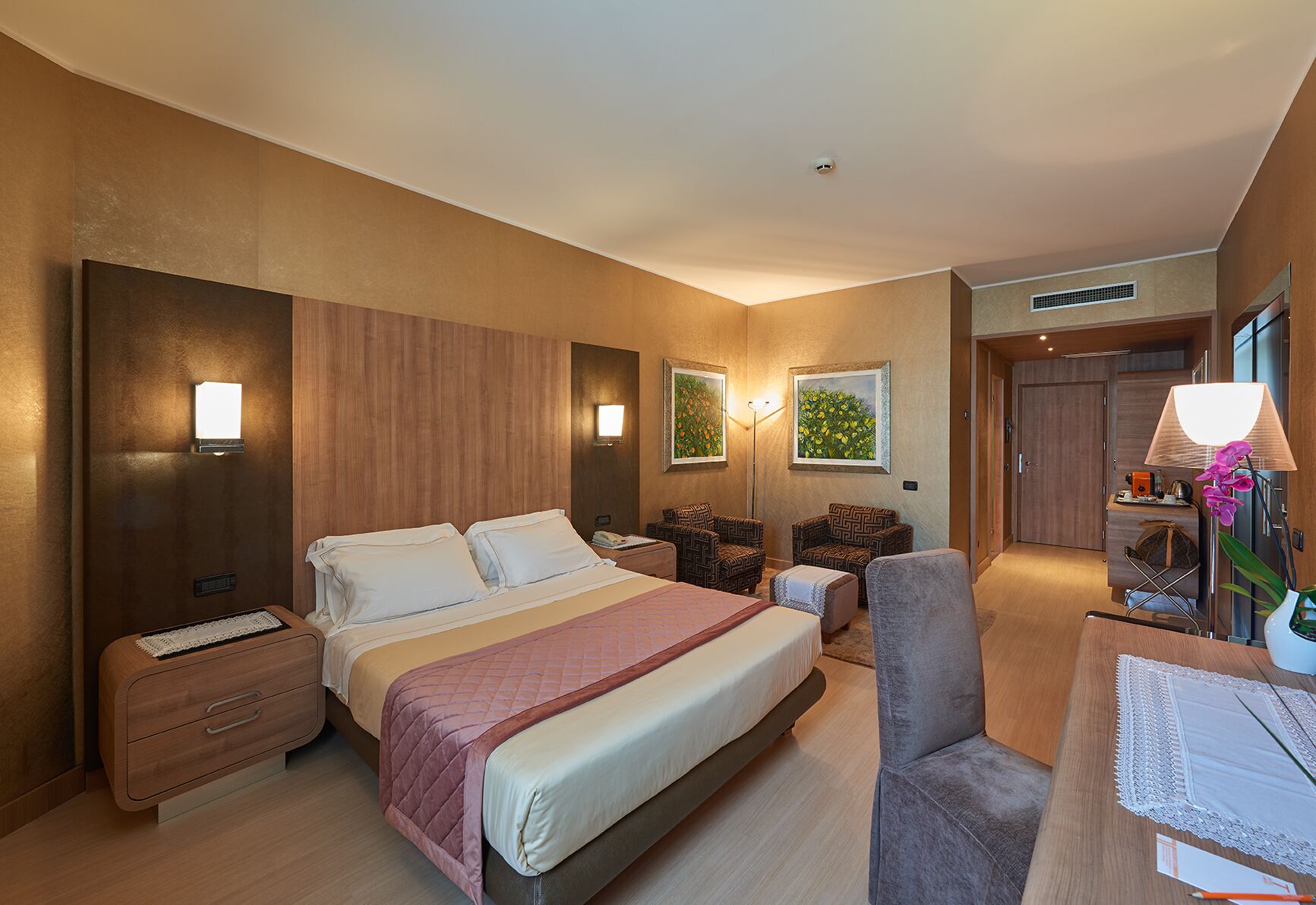 A hotel room with a large bed, two bedside tables, a desk with a chair, and two armchairs. There are wall-mounted lamps and a painting above the desk.