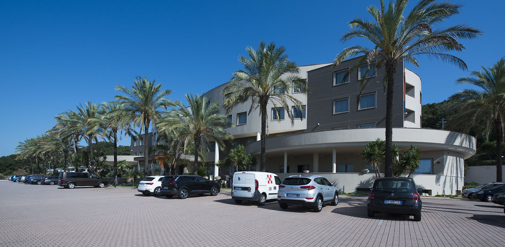 Ein mehrstöckiges Hotel mit geschwungener Fassade, Palmen und einem Parkplatz mit verschiedenen Fahrzeugen.