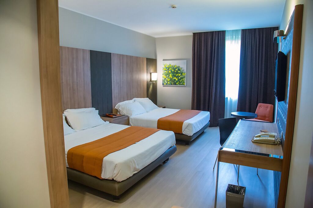 Quarto de hotel com duas camas, uma escrivaninha, uma cadeira e uma janela com cortinas.