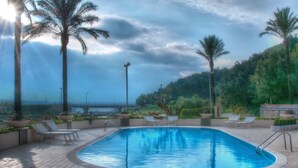 Une piscine entourée de chaises longues et de palmiers, elle-même bordée d’un espace paysager d’où l’on aperçoit un pont au loin.