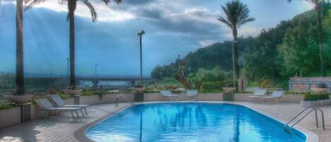 A swimming pool with lounge chairs and palm trees, surrounded by a landscaped area with a view of a bridge in the background.