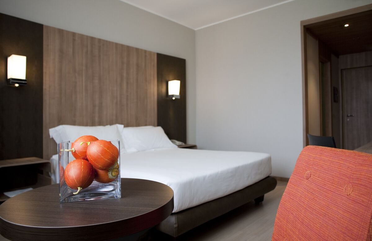 A hotel room with a bed, bedside tables, a round table with a glass vase of oranges, and two wall-mounted lights.