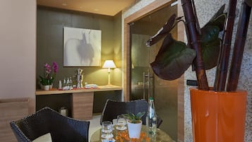 Una camera d'hotel moderna con un tavolo in vetro, sedie in vimini, un divano, una pianta in vaso grande e un'opera d'arte appesa al muro.