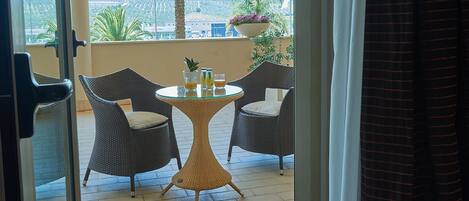 Quarto de hotel com varanda, móveis de vime, uma mesa com bebidas e vista para palmeiras e colinas ao longe.