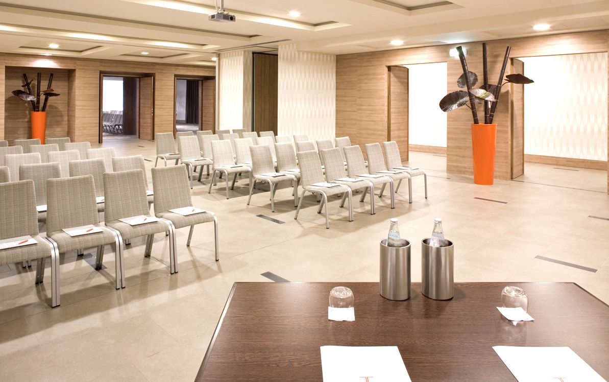 Une salle de conférence avec des rangées de chaises, une table en bois sur laquelle sont posées des bouteilles d’eau et un écran de projection.