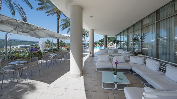 Un espace extérieur aménagé avec des meubles blancs, abrité par un auvent, une piscine en arrière-plan et une vue sur la mer.