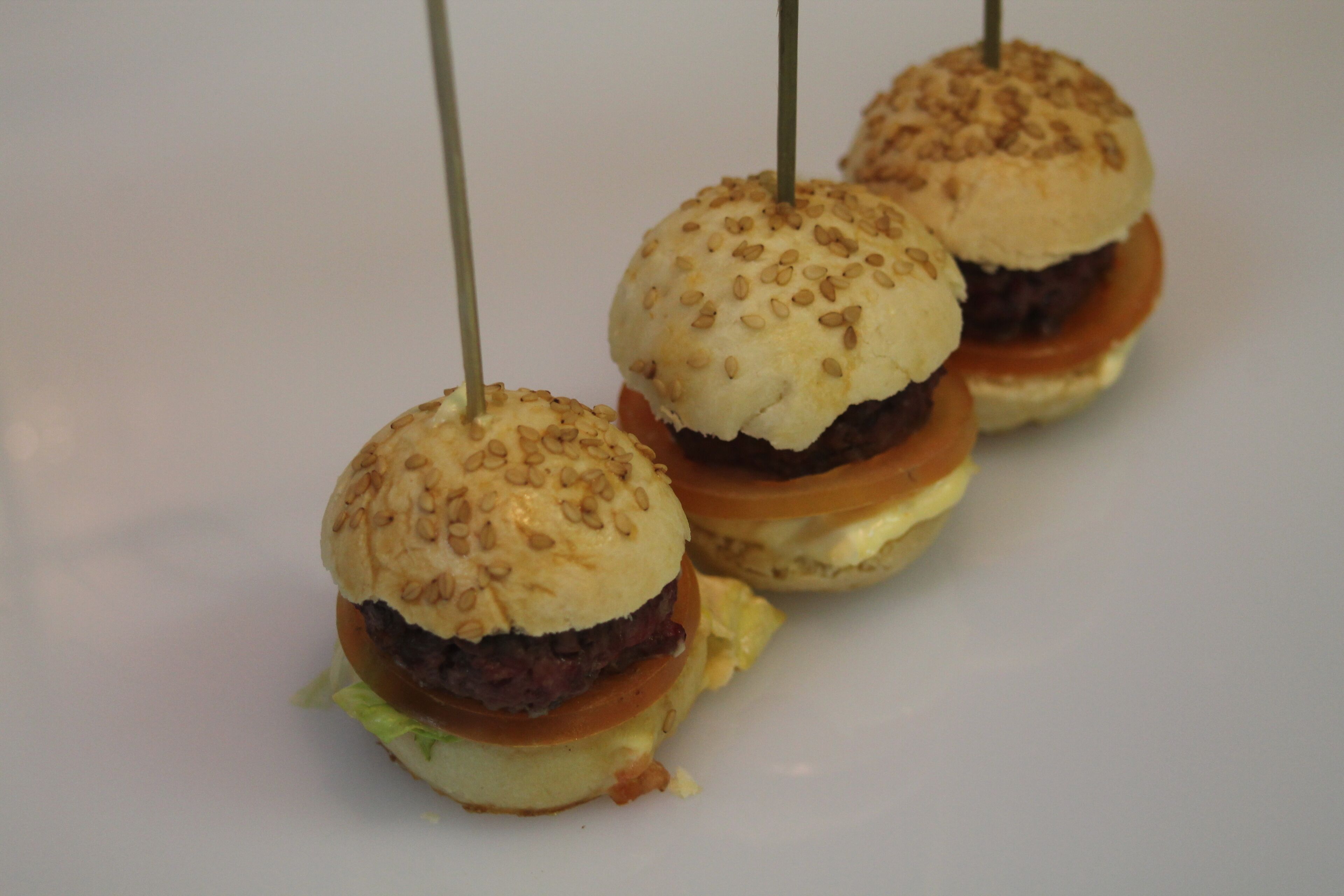Tres hamburguesas pequeñas con panecillos de sésamo y palillos, dispuestas sobre un plato blanco.