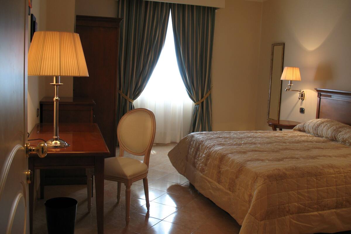 Ein Hotelzimmer mit Bett, Schreibtisch mit Lampe, Stuhl und einem Fenster mit Vorhängen.