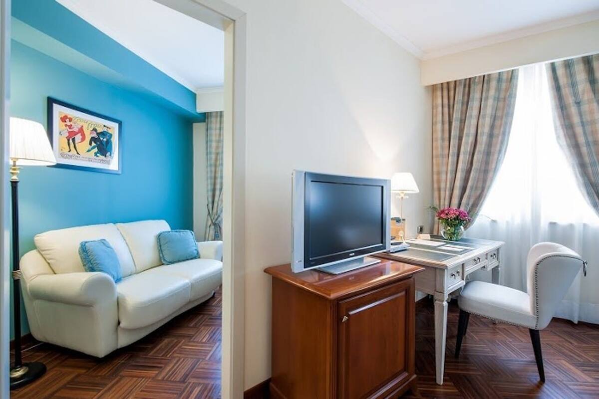 Ein Hotelzimmer mit einer Sitzecke, einem Schreibtisch mit Flachbildfernseher, einem Sessel, einer Lampe und einem Fenster mit Vorhängen.