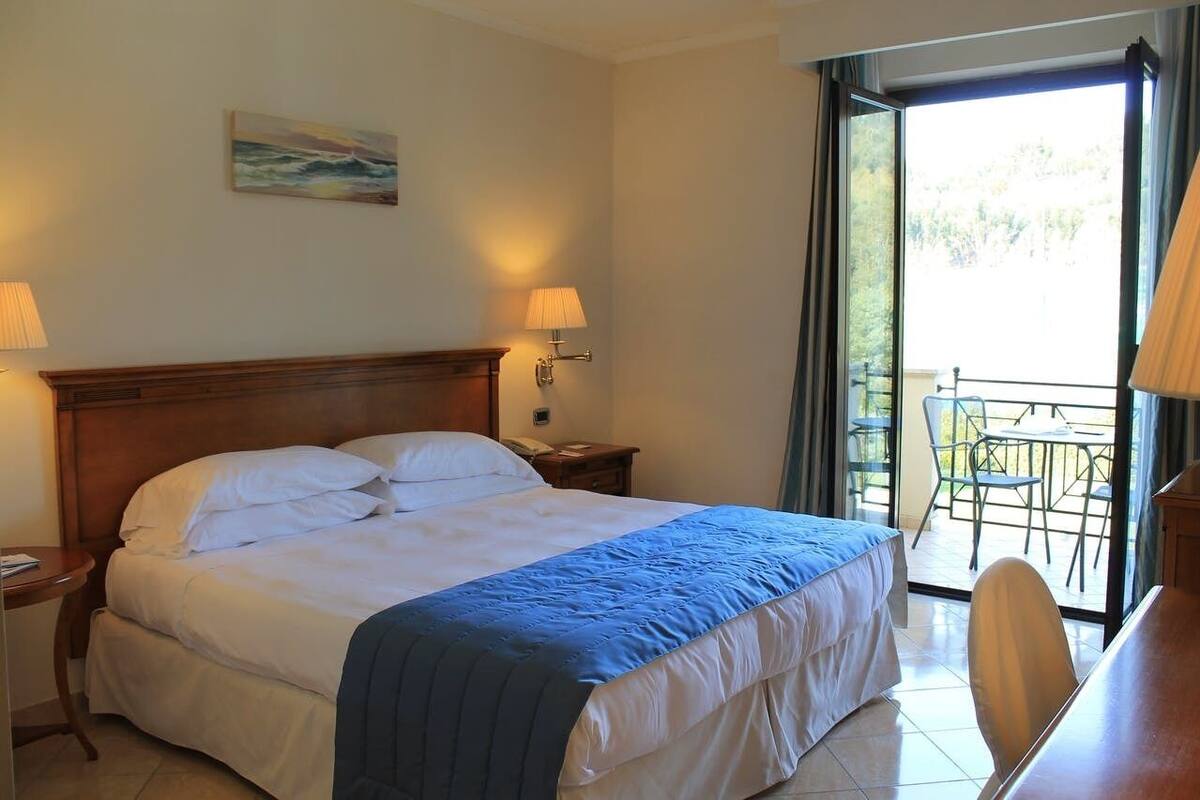 Ein Hotelzimmer mit einem Bett, zwei Nachttischlampen, einem Bild an der Wand und einem Balkon mit Stühlen und einem Tisch.