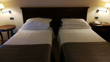 A hotel room with two single beds, each with a pillow and a blanket. There are bedside tables with lamps and a small clock on one side.