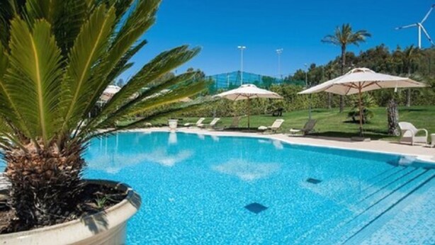Piscina azul cristalina con sillas de descanso y sombrillas, rodeada de follaje y turbinas eólicas al fondo.