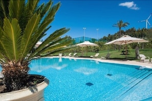 A clear blue pool with lounge chairs and umbrellas, surrounded by greenery and wind turbines in the background. - Hotel San Giorgio (Crotone)