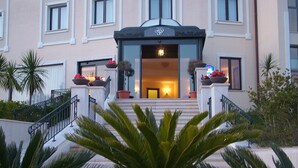 Entrada de hotel con una imponente escalera, un vestíbulo cubierto y plantas en macetas.