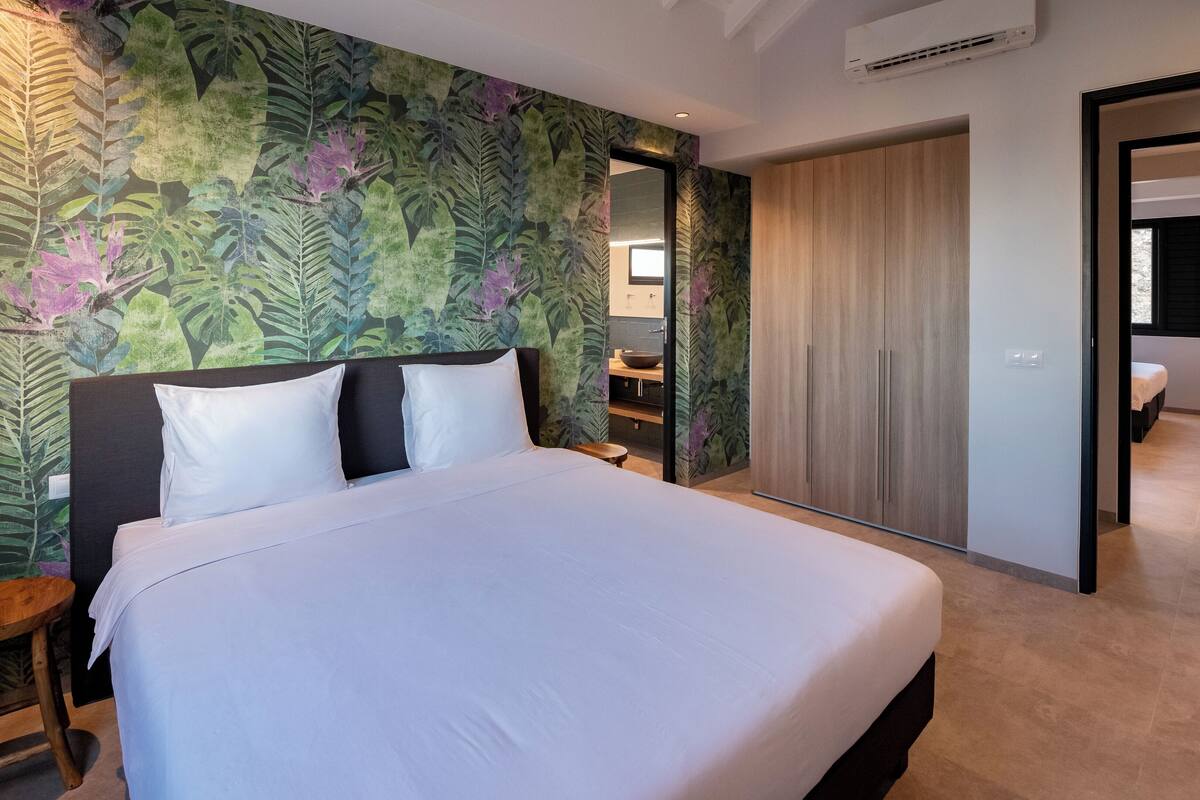 Une chambre d’hôtel avec un lit, une table de chevet en bois, un miroir, une armoire et une salle de bain visible à travers une porte ouverte.