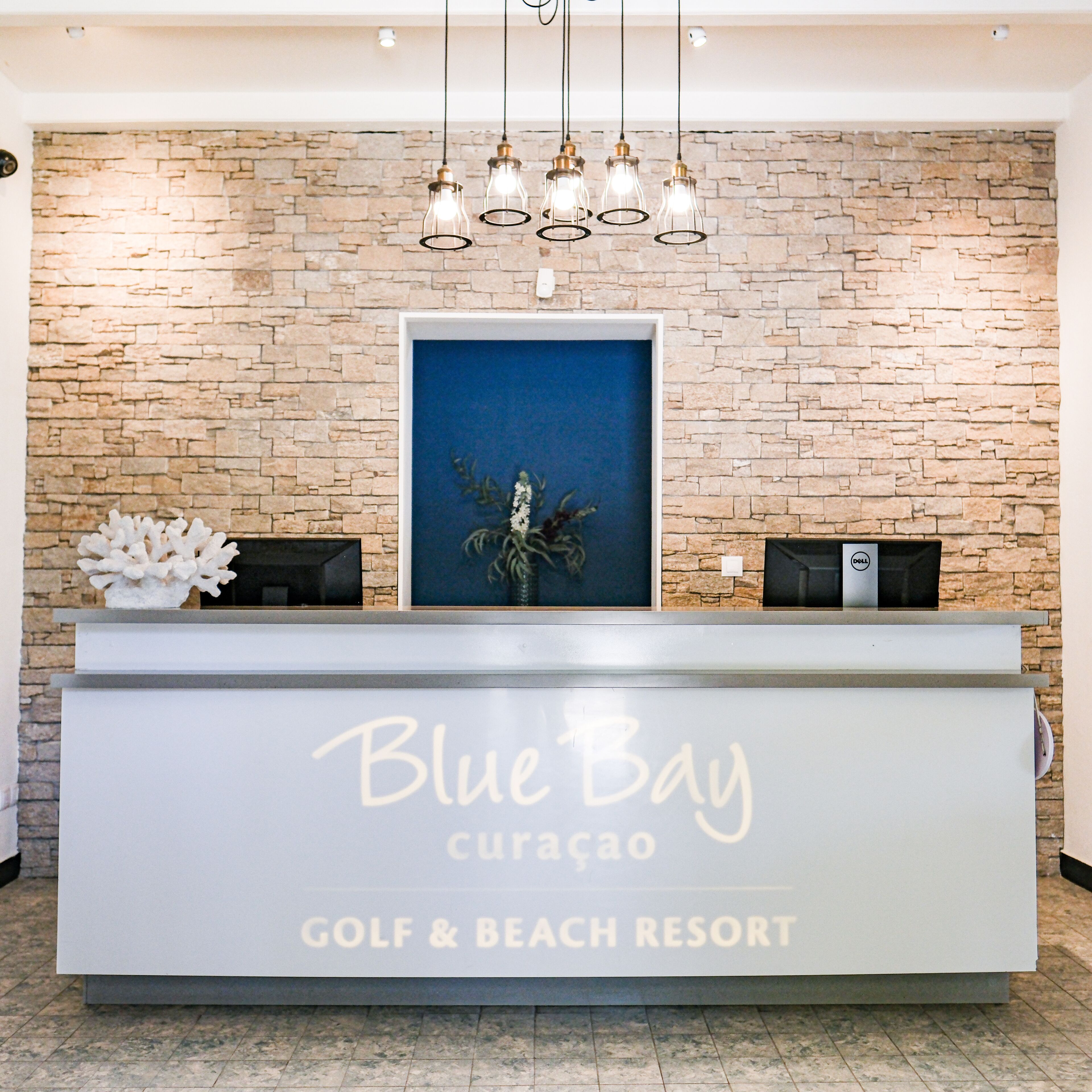 A reception desk at Blue Bay Curacao Golf & Beach Resort with a stone wall background, a framed artwork, and a modern chandelier.