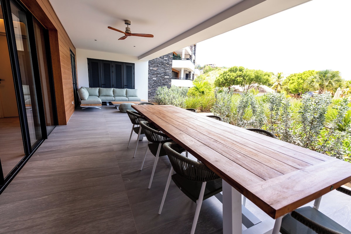 Un espacio moderno para comer al aire libre con una mesa y sillas de madera, un ventilador de techo y vistas a zonas verdes y edificios.