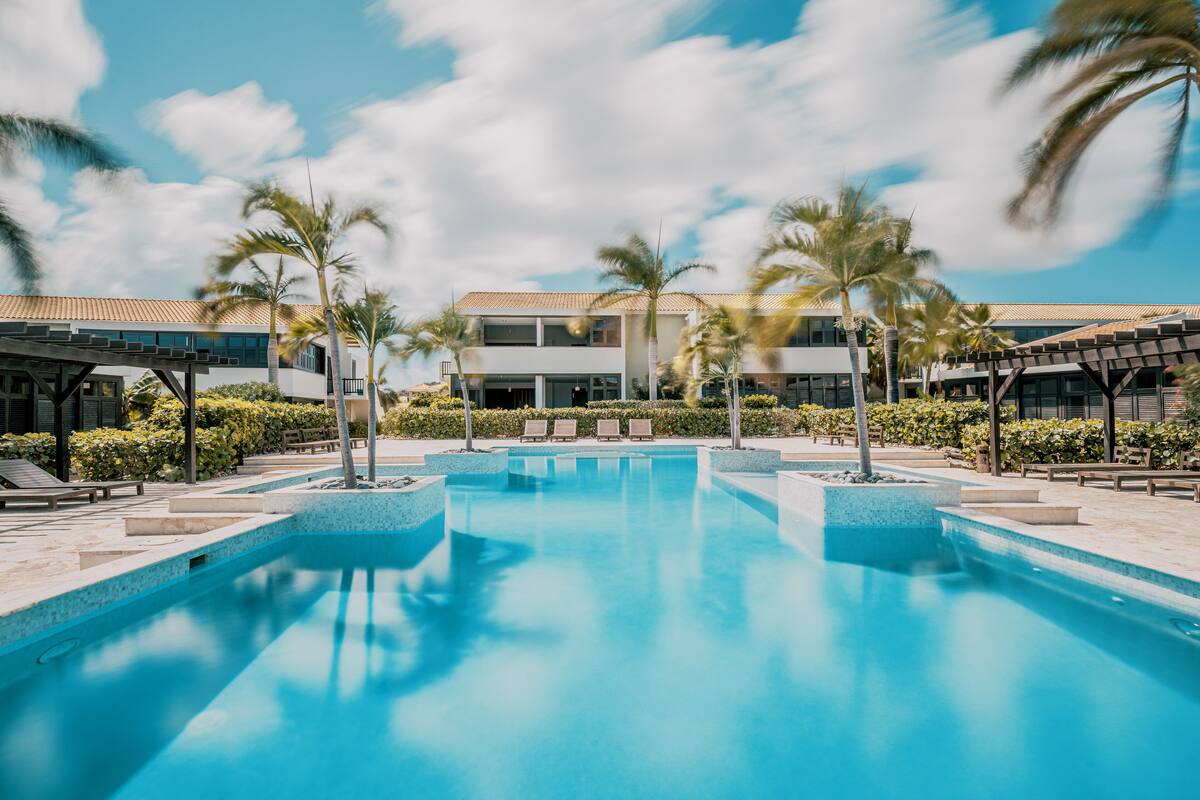 Une piscine entourée de chaises longues et de palmiers, le tout bordé par un bâtiment doté d’un toit en tuiles.