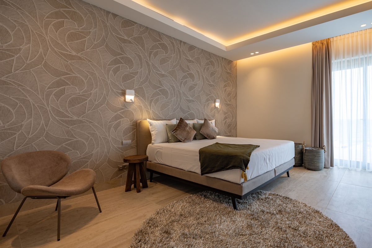 Une chambre à coucher moderne avec un grand lit, une chaise, un petit tabouret et un mur décoratif.