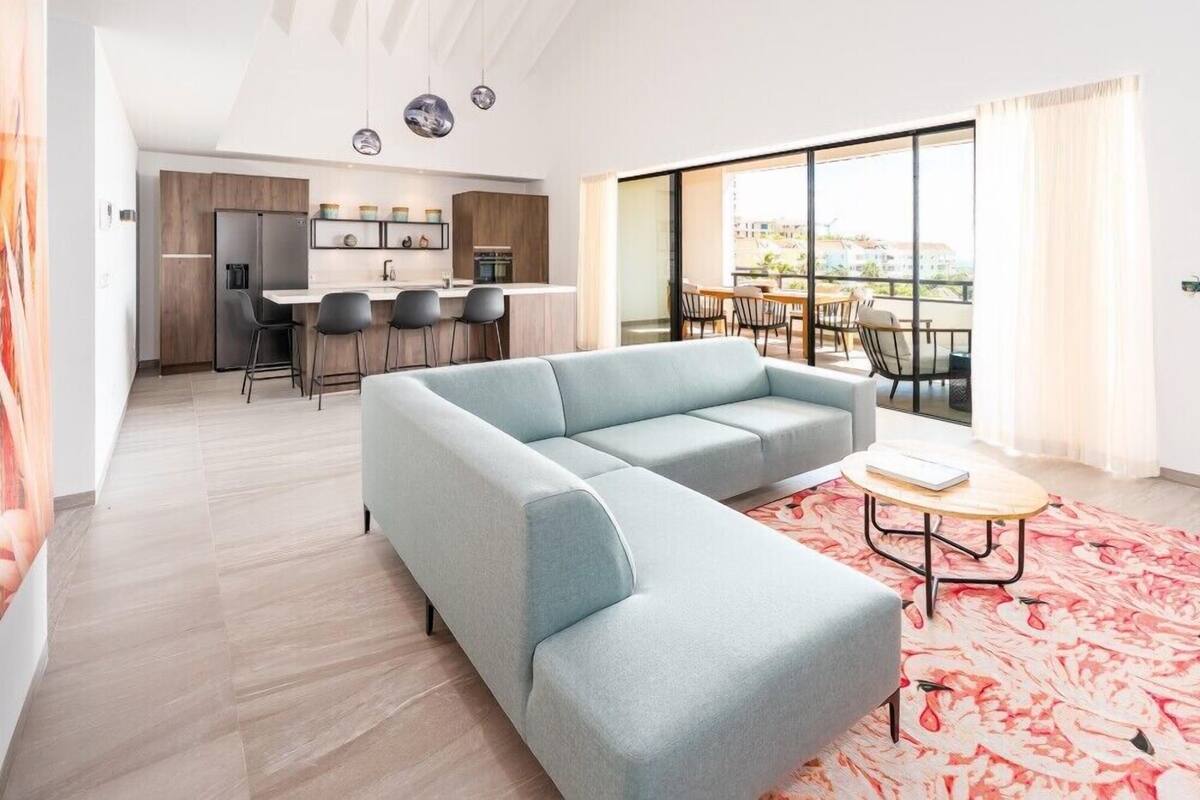 Un salon moderne avec un canapé bleu clair, un coin repas avec une table et des chaises, et une cuisine en arrière-plan.