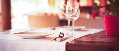 Une table à manger dressée avec une assiette, une fourchette, un couteau et un verre à vin, avec un seau rouge et une plante en arrière-plan.