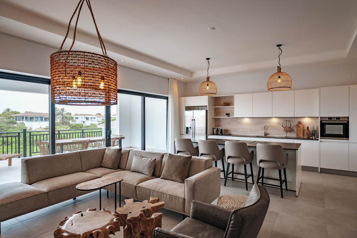 Un salon moderne avec un canapé, une table basse et des fauteuils, une cuisine avec des tabourets de bar et de grandes fenêtres offrant une vue sur l’extérieur.