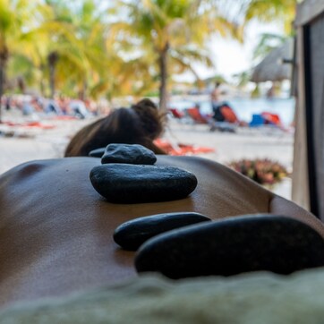 A massage therapy room with a massage table, stones, and a view of a beach with palm trees and lounge chairs.