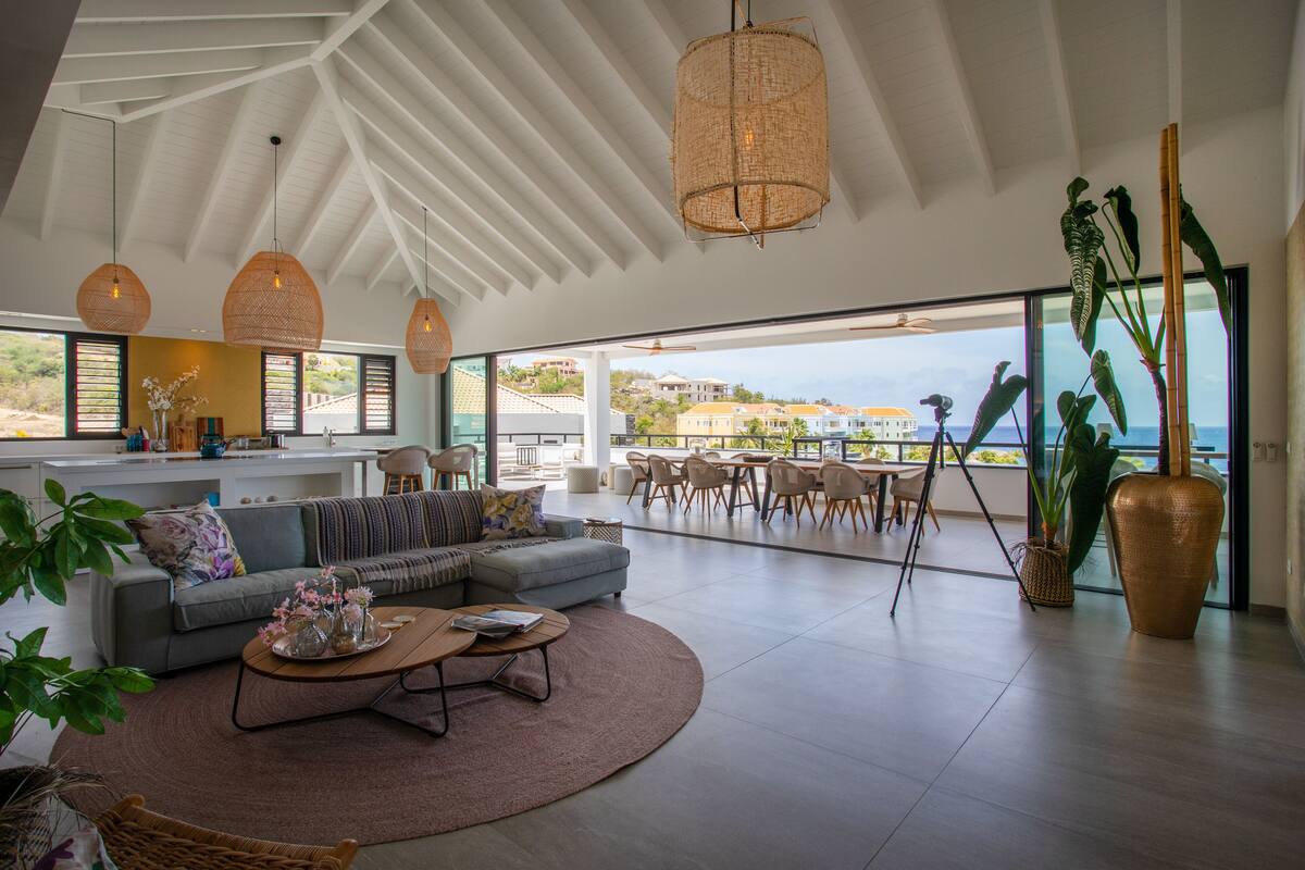 Un vaste espace de vie comprenant un grand canapé, une table basse et un coin repas. On y trouve des lampes en osier et de grandes fenêtres offrant une vue sur la mer.