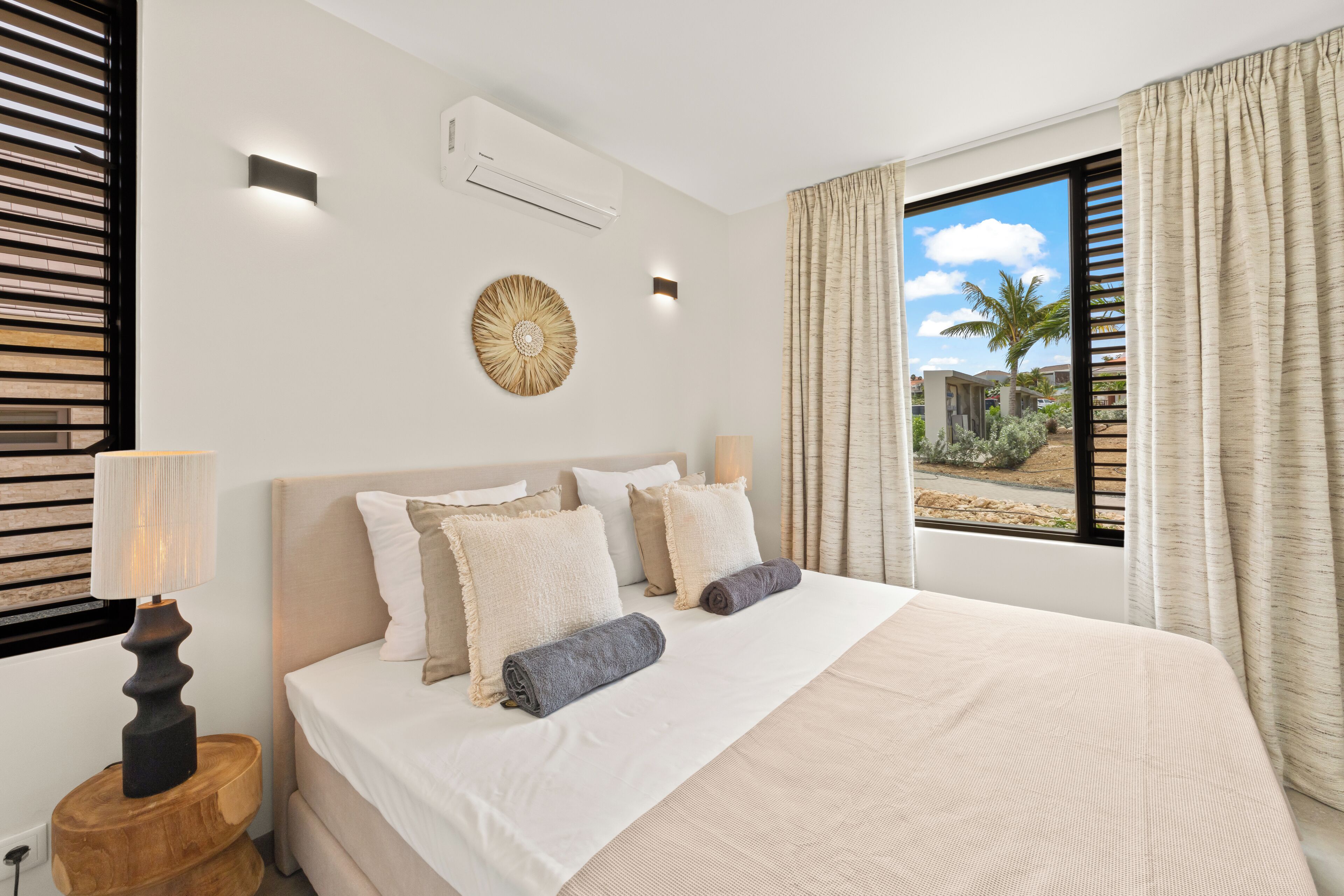 Habitación de hotel con una cama grande, dos lámparas de noche, aire acondicionado instalado en la pared y vista a palmeras a través de la ventana.