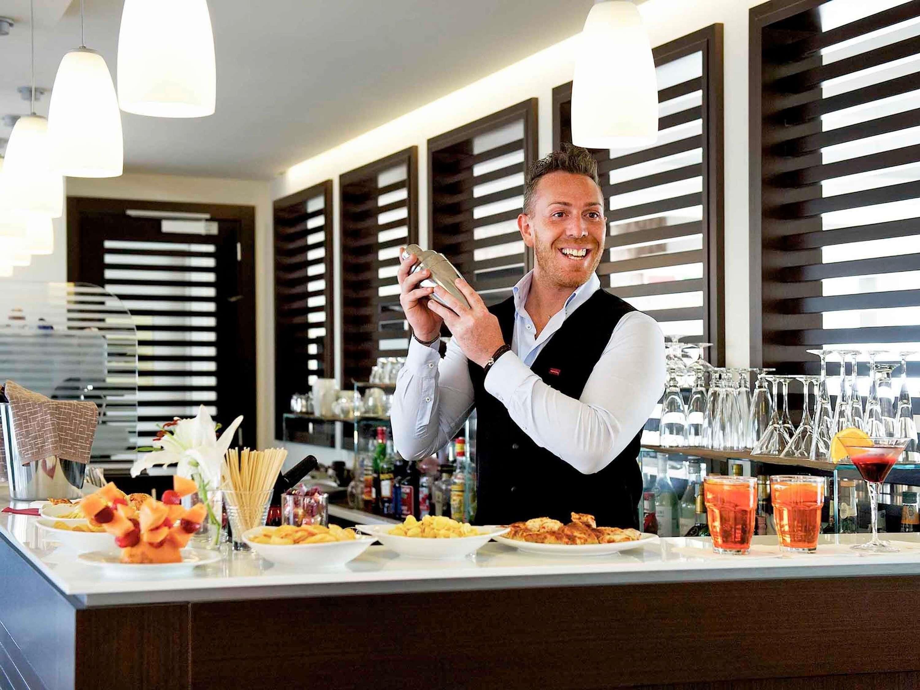 A bar area with a bartender, a counter with plates of food, glasses, and bottles.