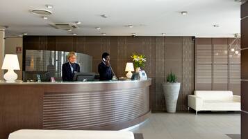 Un mostrador de recepción moderno con una recepcionista, una planta grande en maceta y un sofá blanco en una luminosa zona de vestíbulo.
