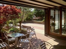Patio esterno con tavolo e sedie in vetro, immerso in un giardino con alberi e un vialetto in mattoni.