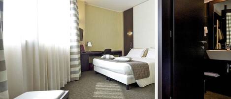 A hotel room with a bed, a desk, a chair, a large mirror, and a bathroom with a sink and toilet.