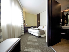 A hotel room with a bed, a desk, a chair, a large mirror, and a bathroom with a sink and toilet.