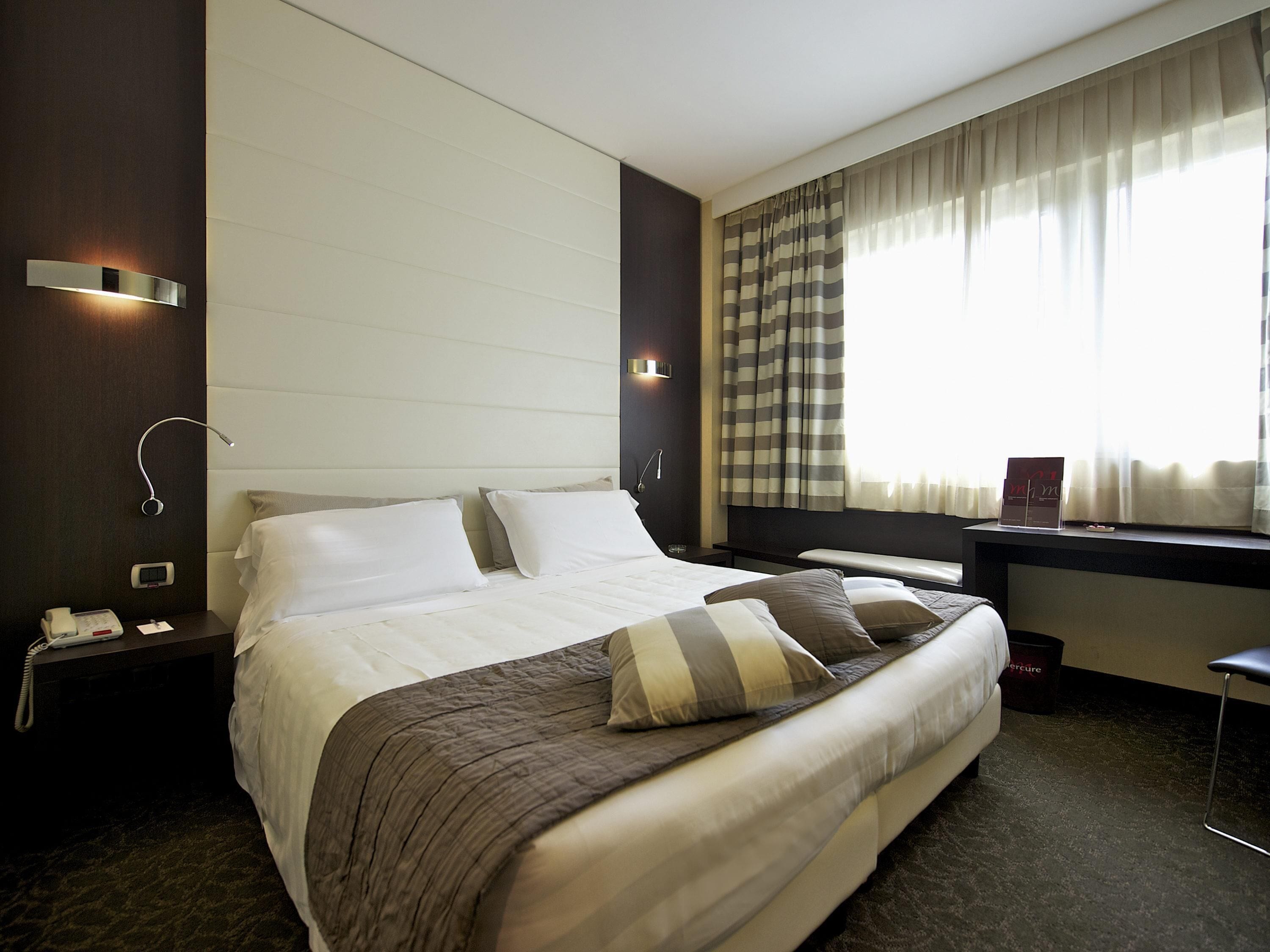 A hotel room with a large bed, bedside tables, a desk, and a window with curtains.
