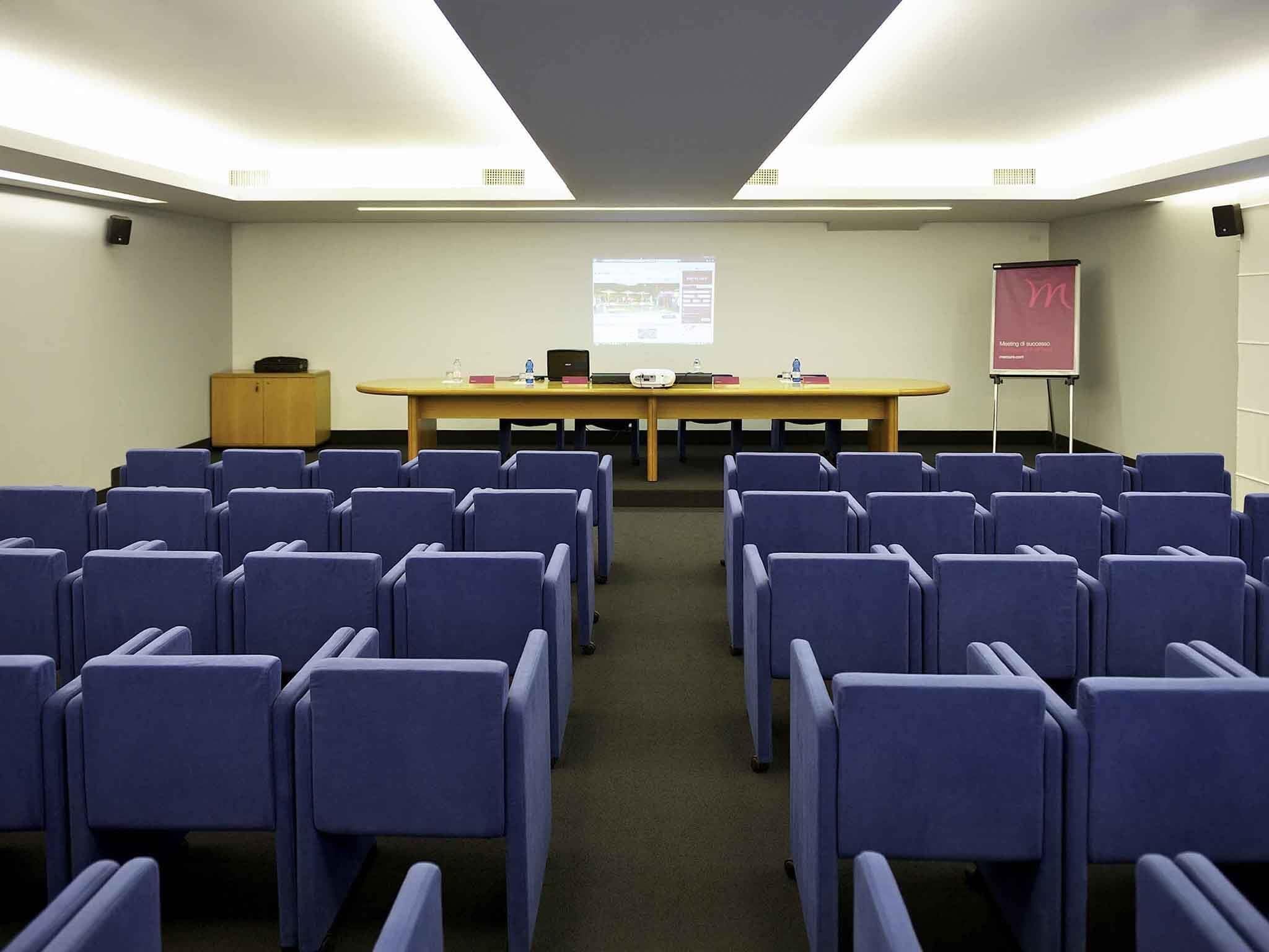 Une salle de conférence avec des rangées de chaises, une longue table, un écran de projection et un pupitre pour les présentations.