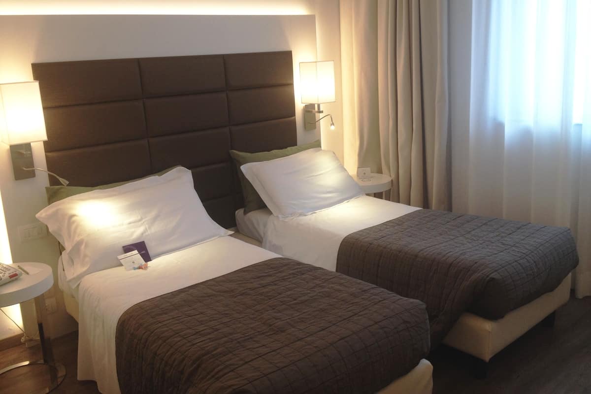 Habitación de hotel con dos camas, cabeceros, lámparas de noche, mesitas de noche y una ventana con cortinas.