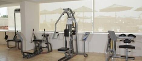 A gym with various exercise machines, large windows with a view of a beach and umbrellas, and a wooden floor.