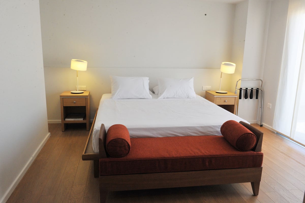 Una cama bien hecha con sábanas blancas, dos mesitas de noche con lámparas, un banco de madera con cojines rojos y un radiador en la pared.