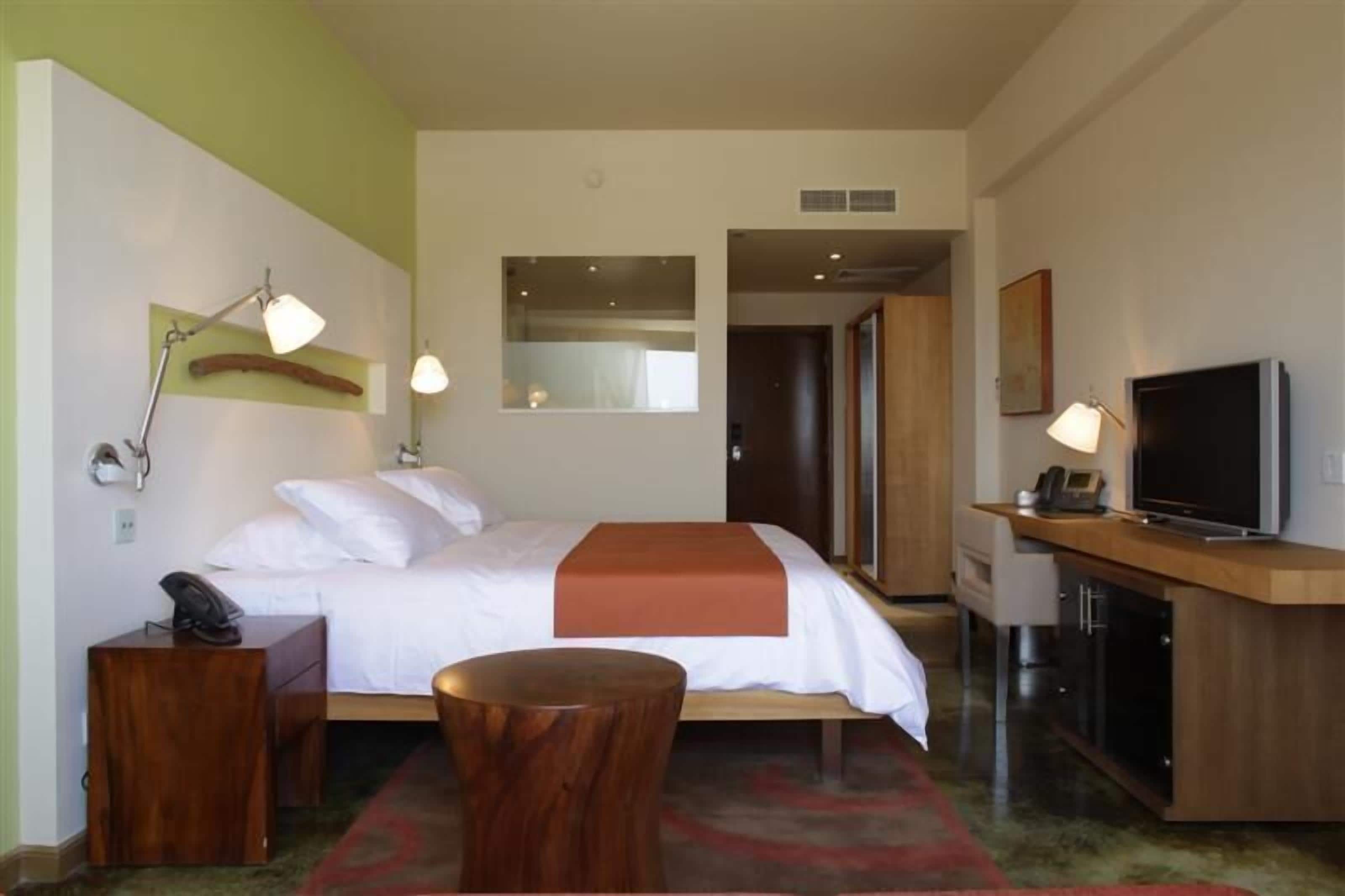 A hotel room with a bed, bedside table, lamp, TV, and a small wooden stool.