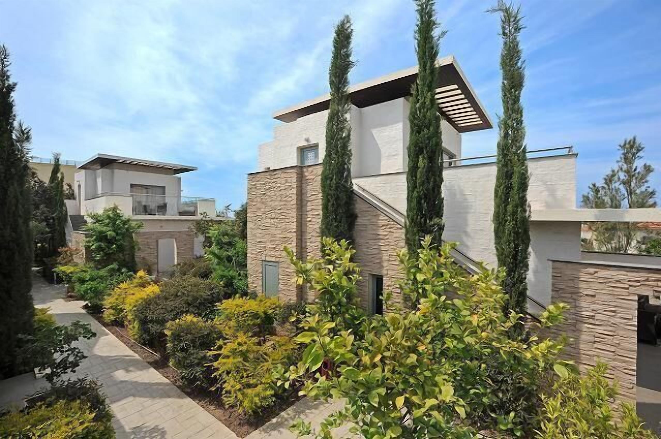 A modern house with a stone facade, a flat roof, and a garden with various shrubs and tall trees.