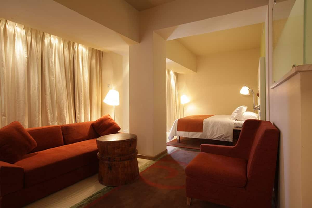 Una habitación de hotel con un sofá rojo, una mesita de madera, una cama con ropa blanca y dos lámparas de noche.