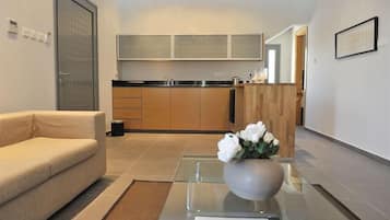 Een moderne woonkamer met een bank, een glazen salontafel met een bloevas, een keuken met houten kasten en een aanrecht, en een deur met een beveiligingspaneel.