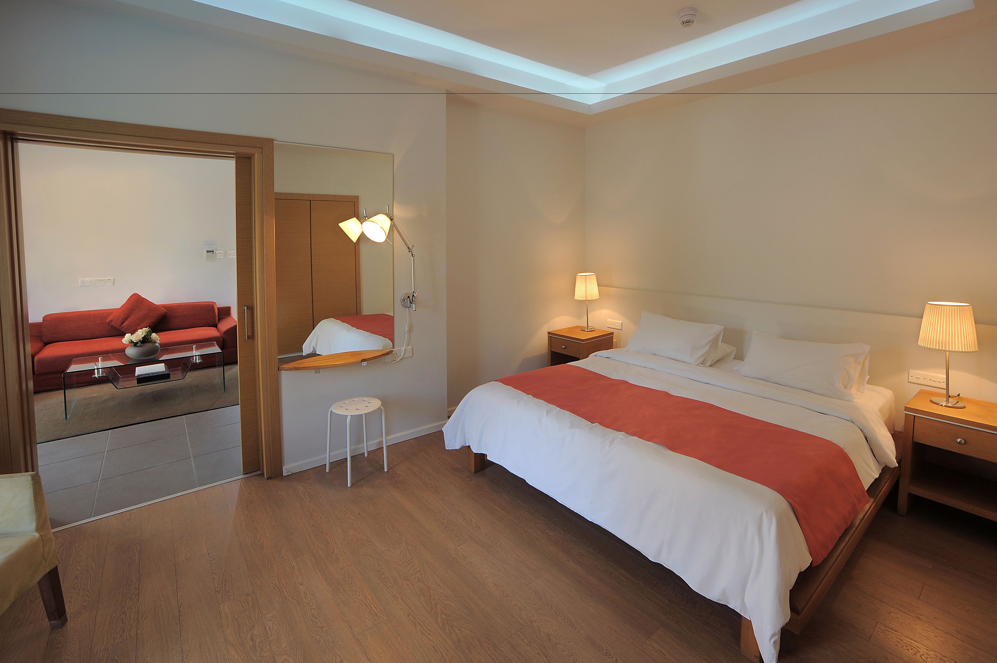 A hotel room with a bed, bedside tables, a small stool, a red sofa, and a mirror.