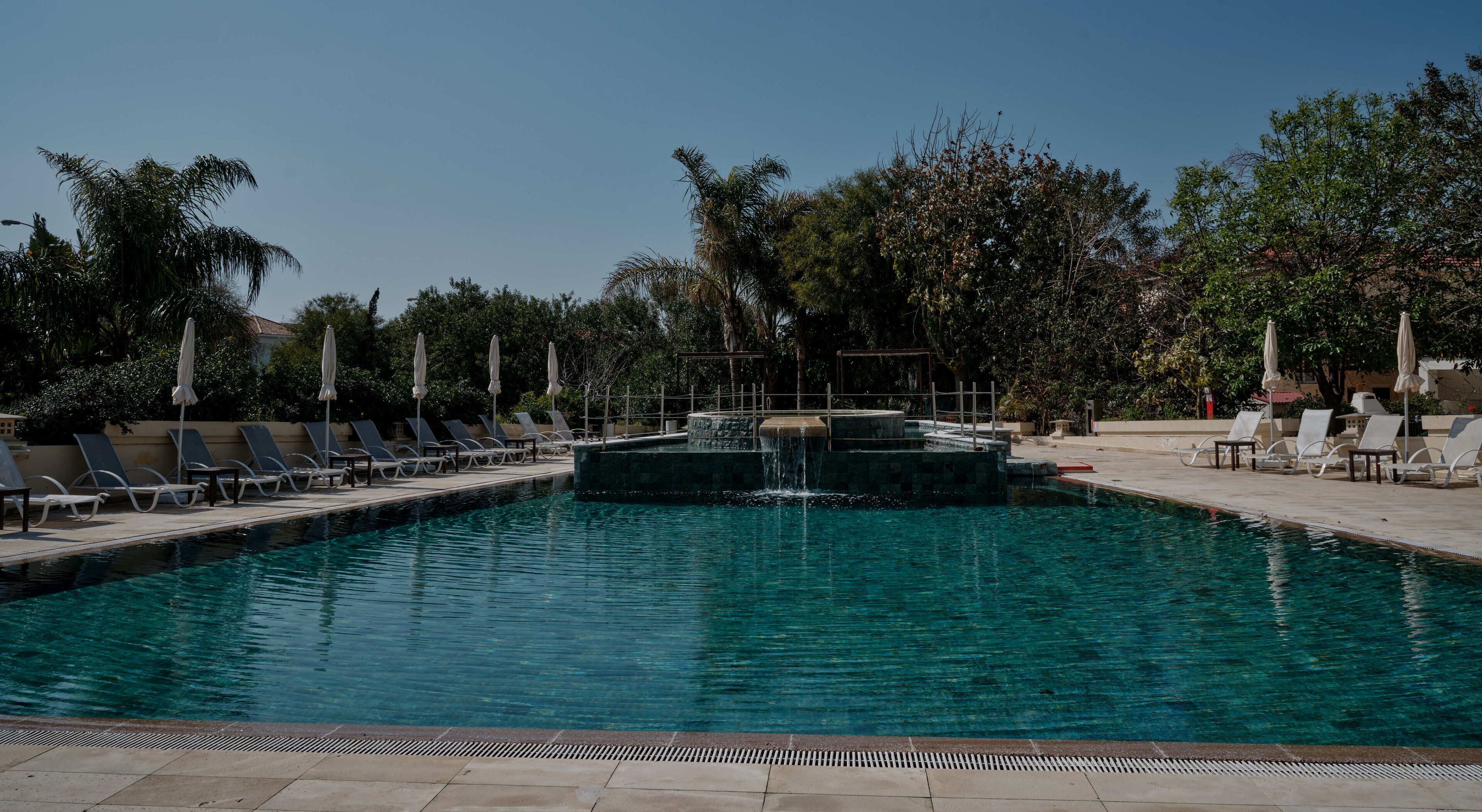 Une piscine entourée d’arbres, avec des chaises longues et des parasols, sous un ciel dégagé.