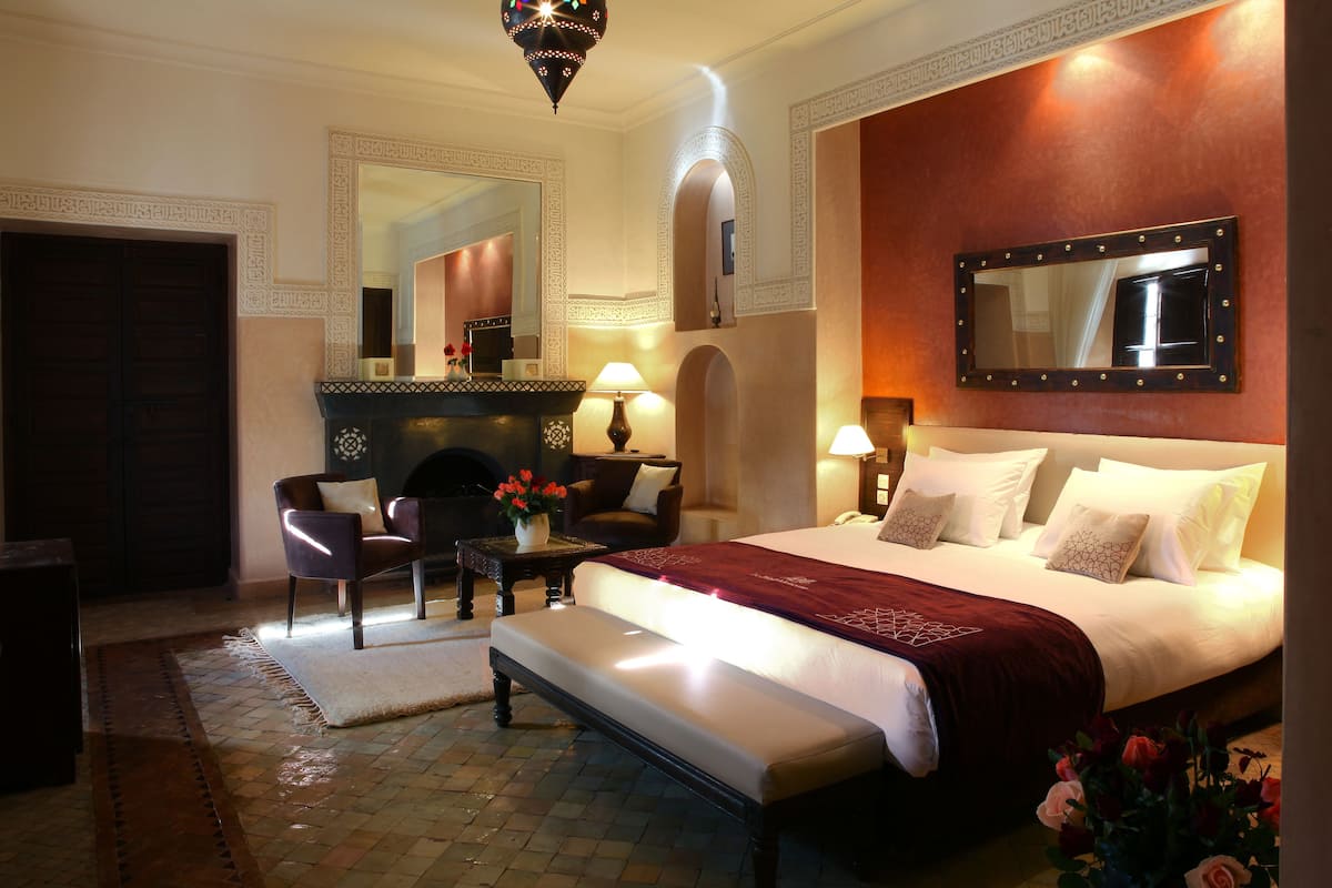 A hotel room with a large bed, a sitting area with a chair and table, a fireplace, and a large mirror on the wall.