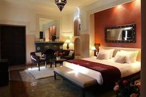 A hotel room with a large bed, a sitting area with a chair and table, a fireplace, and a large mirror on the wall.